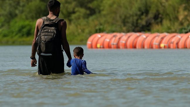 FILE - Migrants walk past large buoys being used as a floating border barrier on the Rio Grande, Aug. 1, 2023, in Eagle Pass, Texas. (AP Photo/Eric Gay, file)