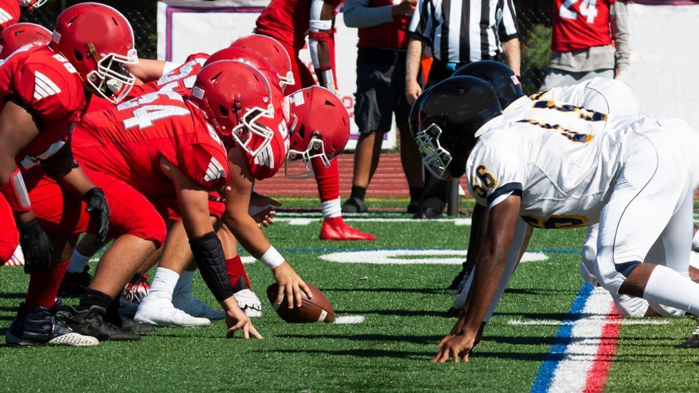 Side view of two high school football teams lined up for the snap of the ball on a green turf field.