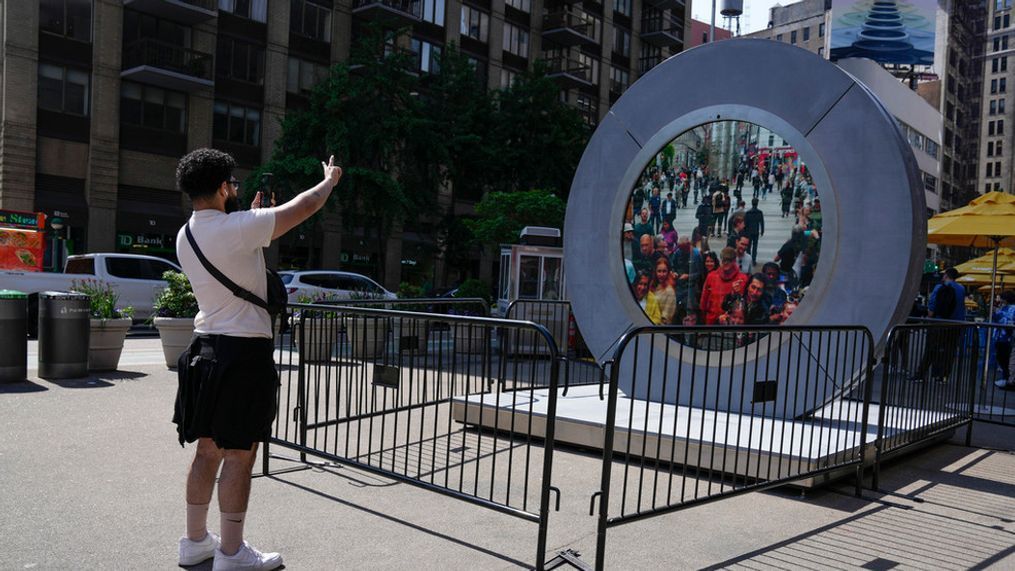 A man signals to pedestrians in Dublin, Ireland through a livestream portal as part of an art installation on the street in New York, Tuesday, May 14, 2024. (AP Photo/Seth Wenig)