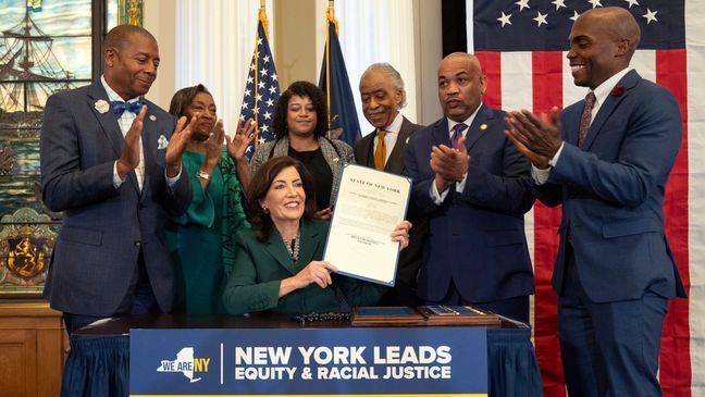 This image provided by the Office of The Governor, shows New York Gov. Kathy Hochul, seated center, after she signed a bill, in Albany, NY, Tuesday, Dec. 19, 2023, to create a commission tasked with considering reparations to address the persistent, harmful effects of slavery in the state. (Don Pollard/Office of the Governor via AP)